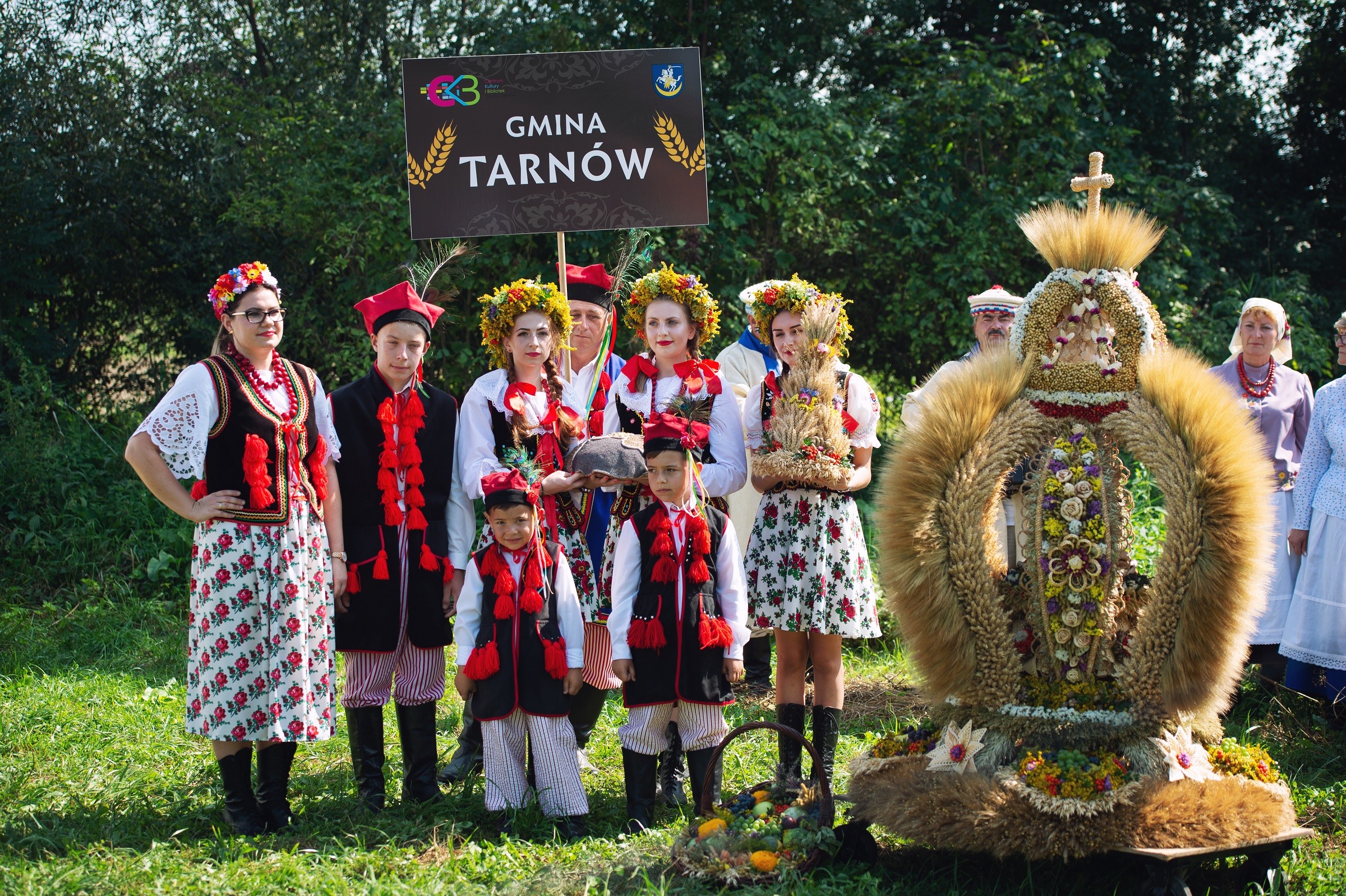 grupka osób w strojach ludowych stoi obok wysokiego wieńca zbożowego zwieńczonego zbożowym krzyżem